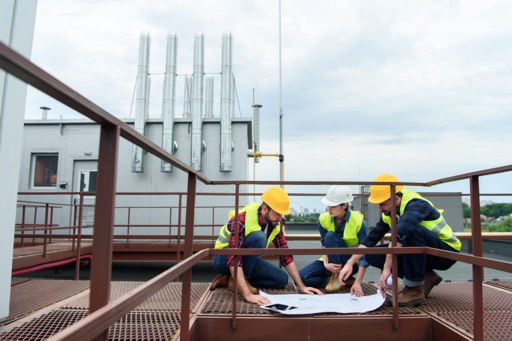 three-engineers-in-helmets-working-with-blueprints-on-roof-together